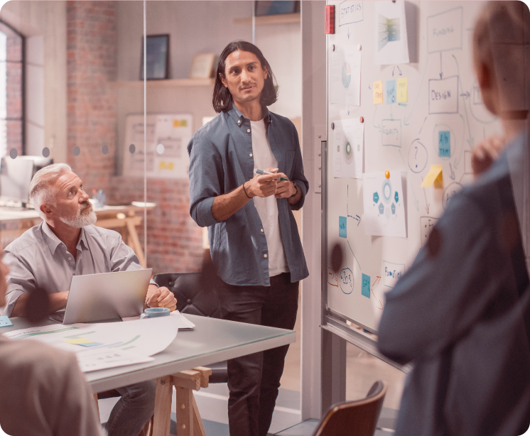 A team meeting in a modern office, with a young man standing and presenting at a whiteboard to colleagues.